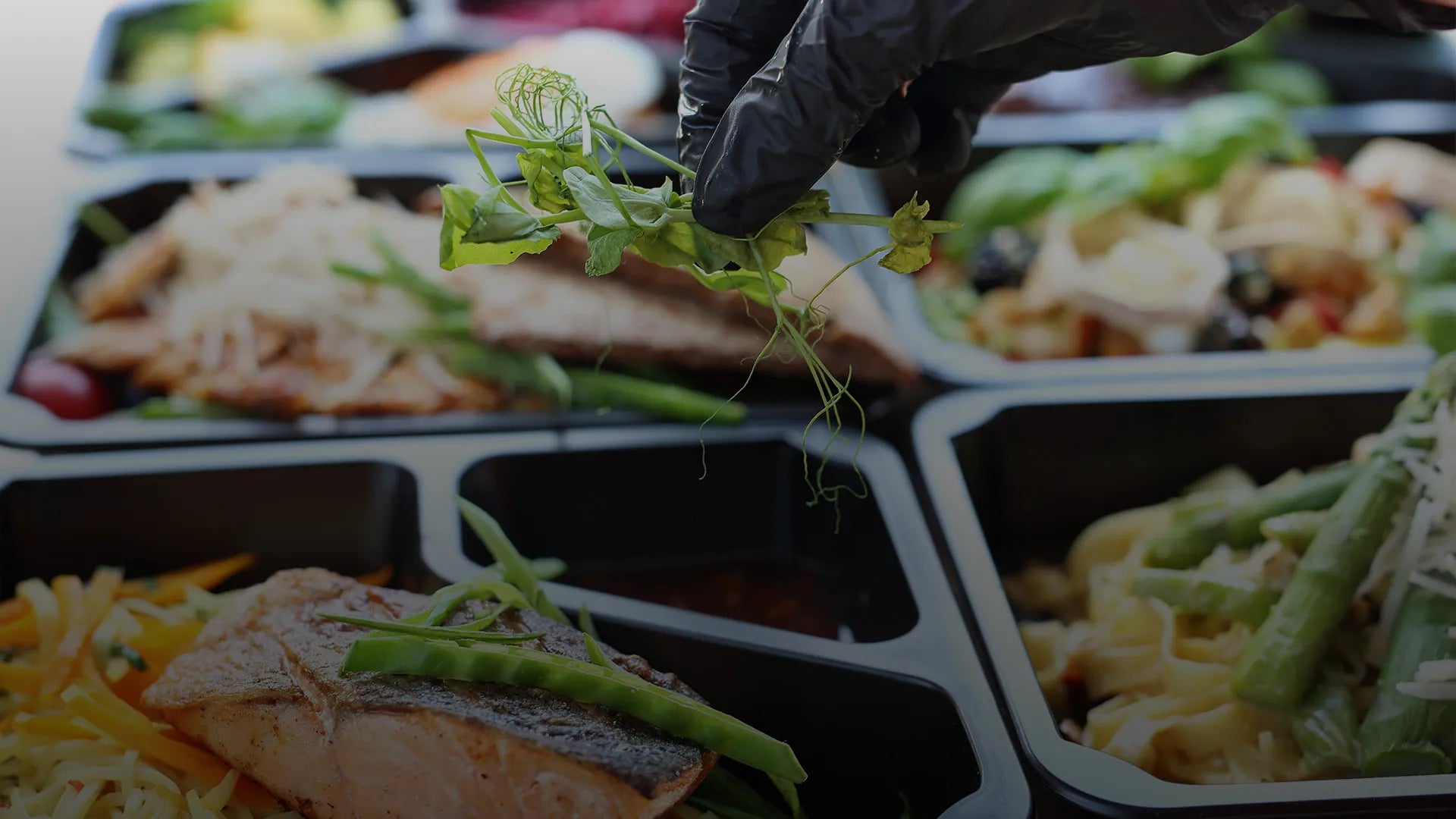 Hand in a black glove arranging greens on a salad in a compartmentalized food container.